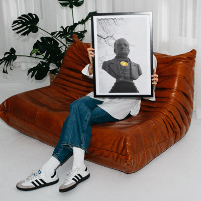 A woman sitting comfortably on a contemporary brown sofa, proudly holding a framed sunset print. This image showcases stylish home decor featuring canvas prints UK that brighten any living space.